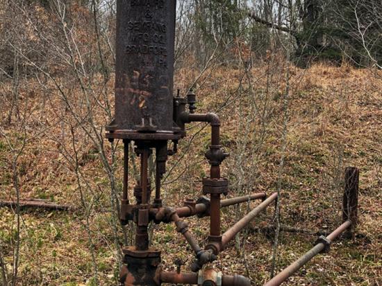 An orphan well with rusted equipment above it surrounded by brush