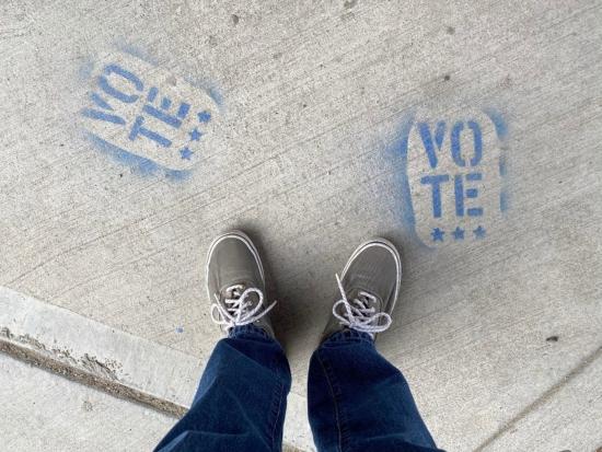 Person in jeans and grey sneakers standing on sidewalk that has been painted with the word "Vote"