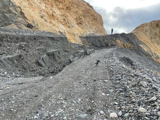 A gravel road on a mountainside, with an enormous fracture causing a big drop in the road bed