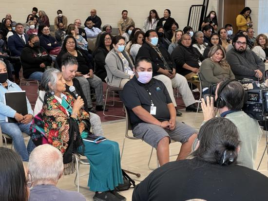 Large room with crowd seated in rows of chair. At the front of the crowd a woman holds a microphone. 