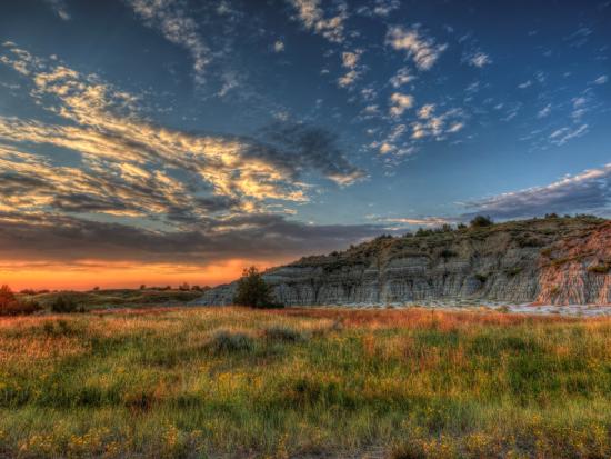 Green prairie grasses in the foreground with a butte in the background and cloudy sky above. 