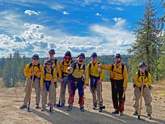 Wildland firefighters stand in a row with a forested landscape in the background. 
