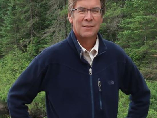 Image of Robert Anderson; he is a light-skinned middle aged man with brown hair and a blue zipped sweatshirt in the woods.