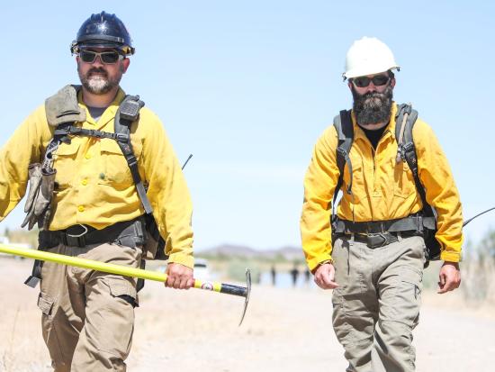 Two wildland firefighters walk towards the viewer wearing their gear. 