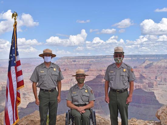 Three park rangers stand at attention beside the American flag in front of the Grand Canyon