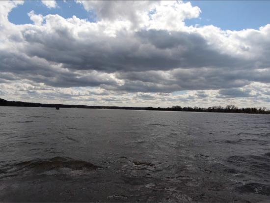 A river flows past a distant shoreline under a sky with clouds 
