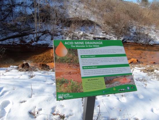 Discolored water flows through a stream near abandoned coal mines, a sign saying “Acid Mine Drainage” marks the spot