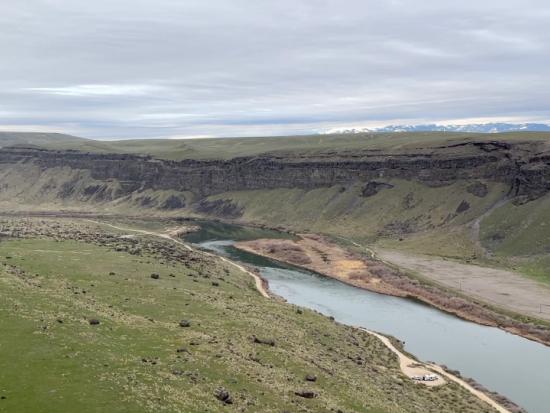 A river flows between two high grassy bluffs, with snow-covered mountains in the distance