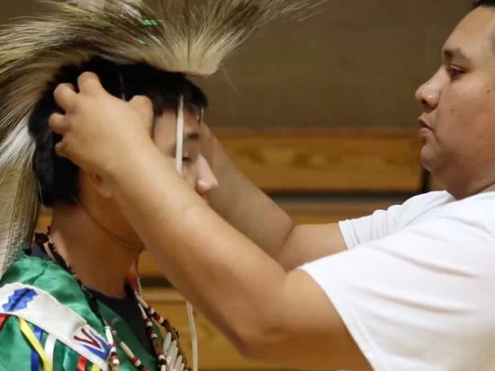 An Indigenous man adjusts a younger man’s ceremonial headdress.