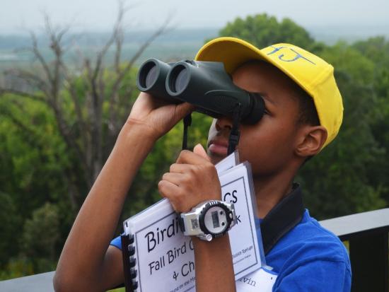 Child with binoculars, wearing a yellow hat and holding a book about birds.