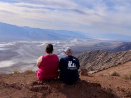 Two people sit on the ground with their back to the camera. A broad white and brown valley is beyond them.