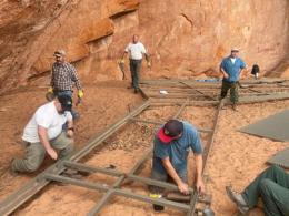 Five workers building a wooden walkway on red earth. 