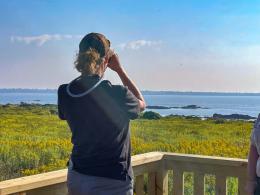 Martha Williams standing on a wooden platform looks away from the camera through binoculars.