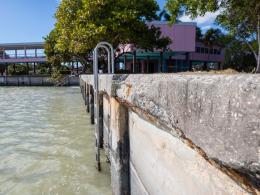 Deteriorating edge of seawall next to opaque light green water and a pink building in the background