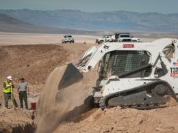 White excavator digs dirt from ditch in Death Valley National Park with construction team beside it