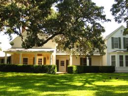 A two-story white frame and stone house is shaded by a large tree.