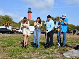 People with shovels break ground with tall, red lighthouse in the background.