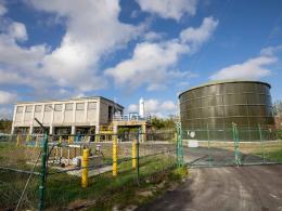 Large water tank, elevated concrete building and outdoor pipes behind a fence.