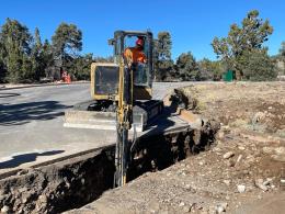 A worker in an orange safety vest and red hardhat is inside the cab of a small excavator, operating the bucket to dig a trench through a parking lot. The trench will be used for water and wastewater lines.