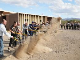 A line of people holding shovels with yellow handles throw a cloud of brown dirt while people watch.