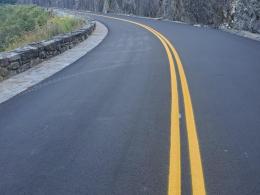 Freshly paved mountain road with double yellow lines curving beside a stone wall and steep, tree-covered rock face