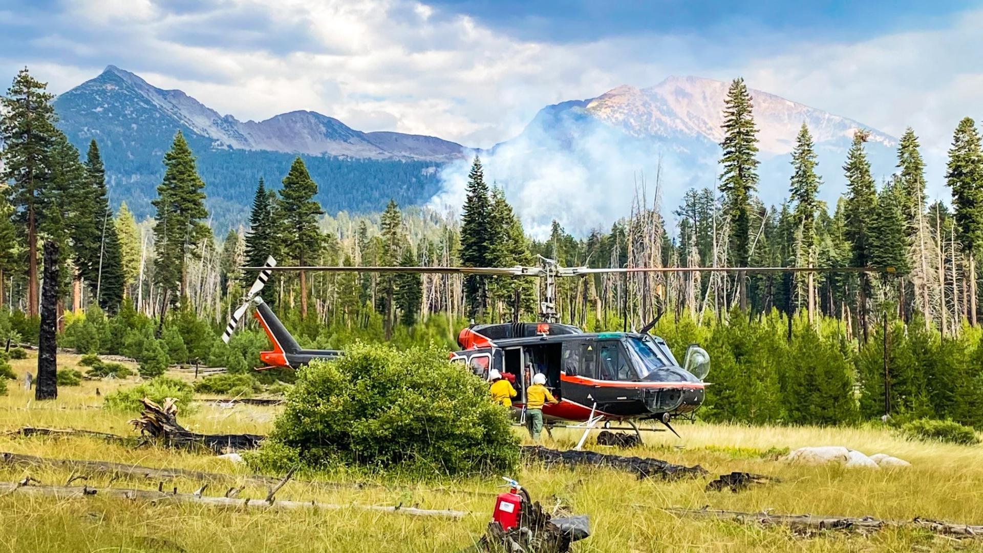 Members of a Yosemite helitack crew work around a helicopter in a grassy field. Trees border the field, with smoke rising from their midst. Mountains rise in the distance under a stormy sky. Photo by Yosemite National Park.
