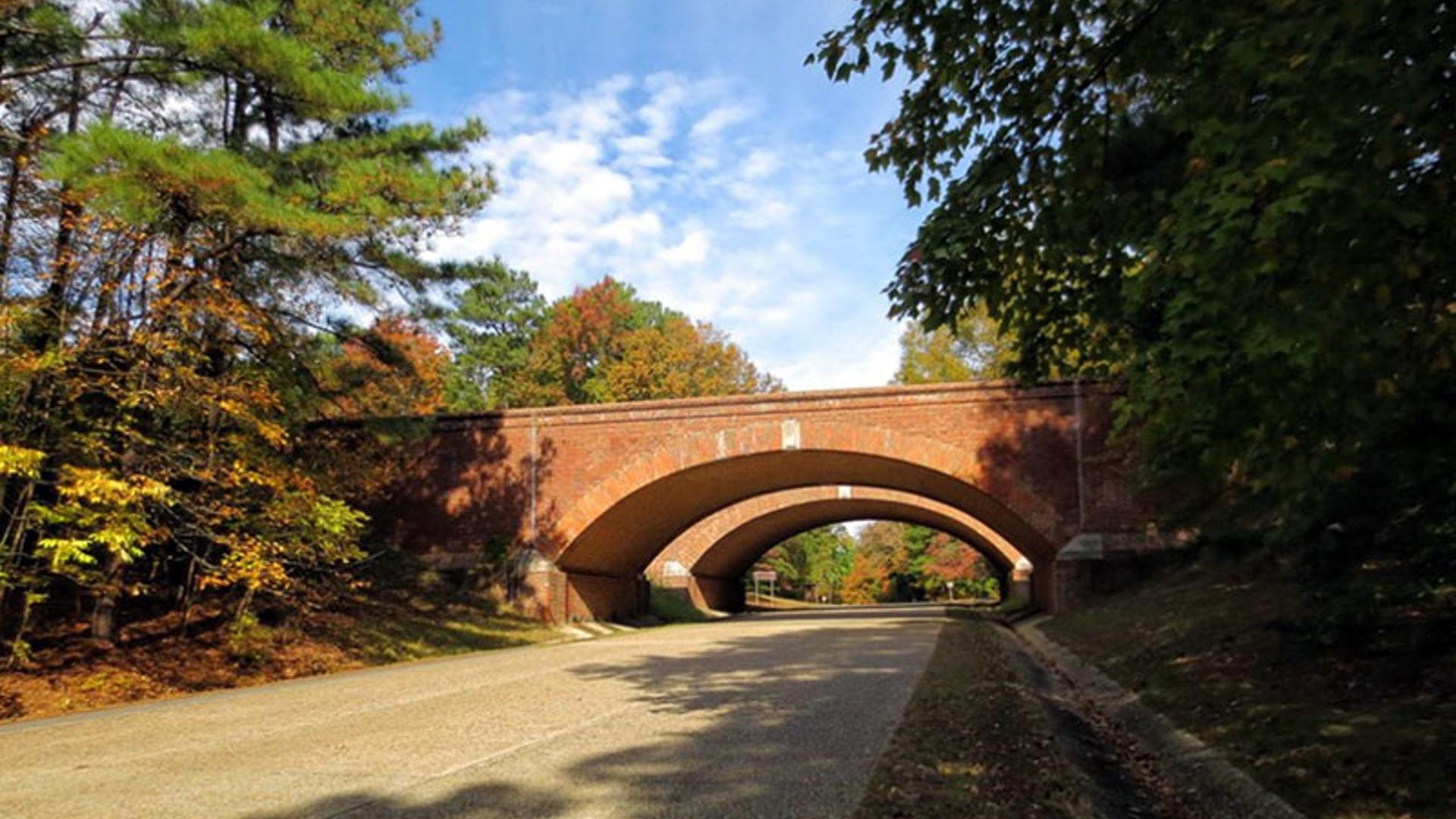 Paved road leading to two red stone bridges with trees on either side and a blue sky
