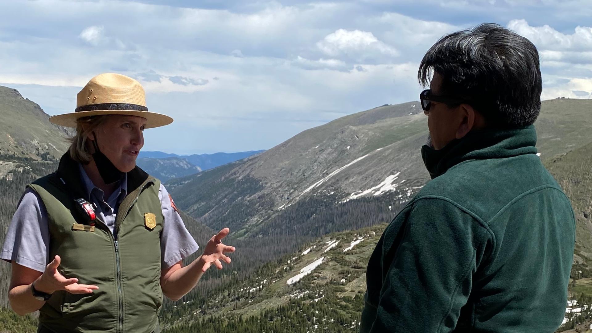 Man in green jacket speaks with park ranger with mountain scenery and blue sky in background.