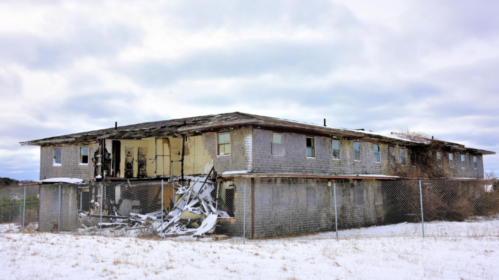 A large building sits behind fencing with walls torn away, exposing the interior of the building