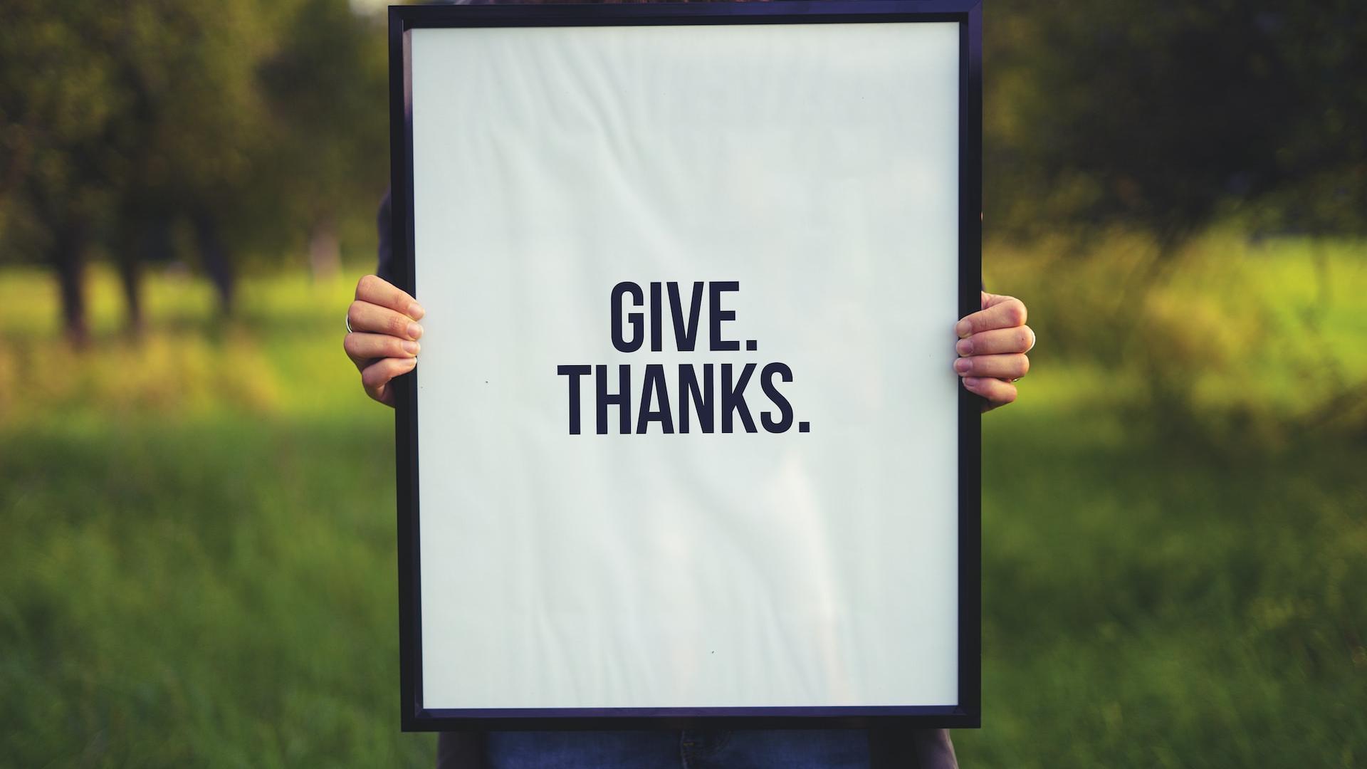 Person holding a sign in a field that says "Give. Thanks."