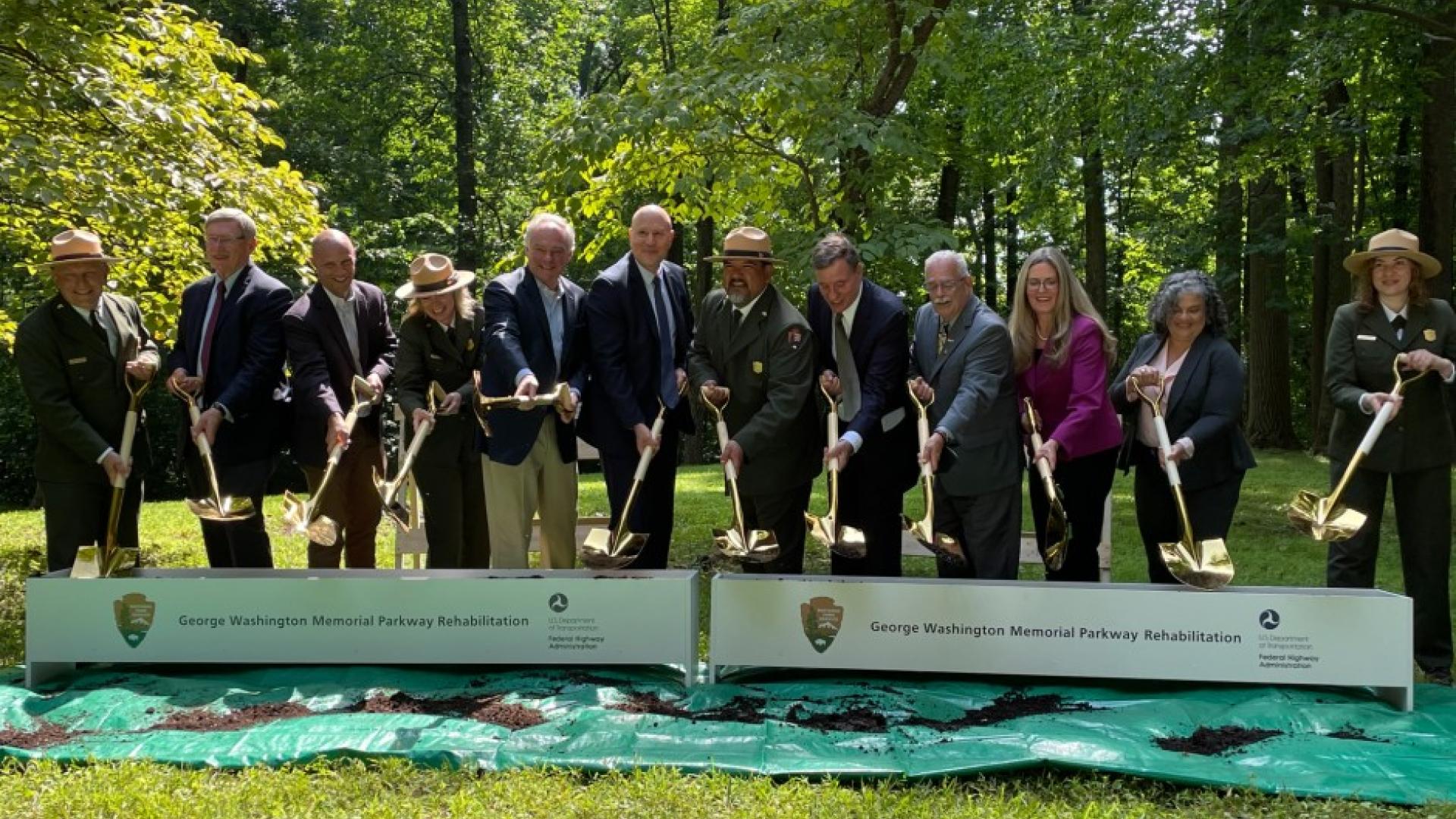 Various people, wearing suits and green uniforms, dig dirt behind a banner reading "George Washington Memorial Parkway Rehabilitation".