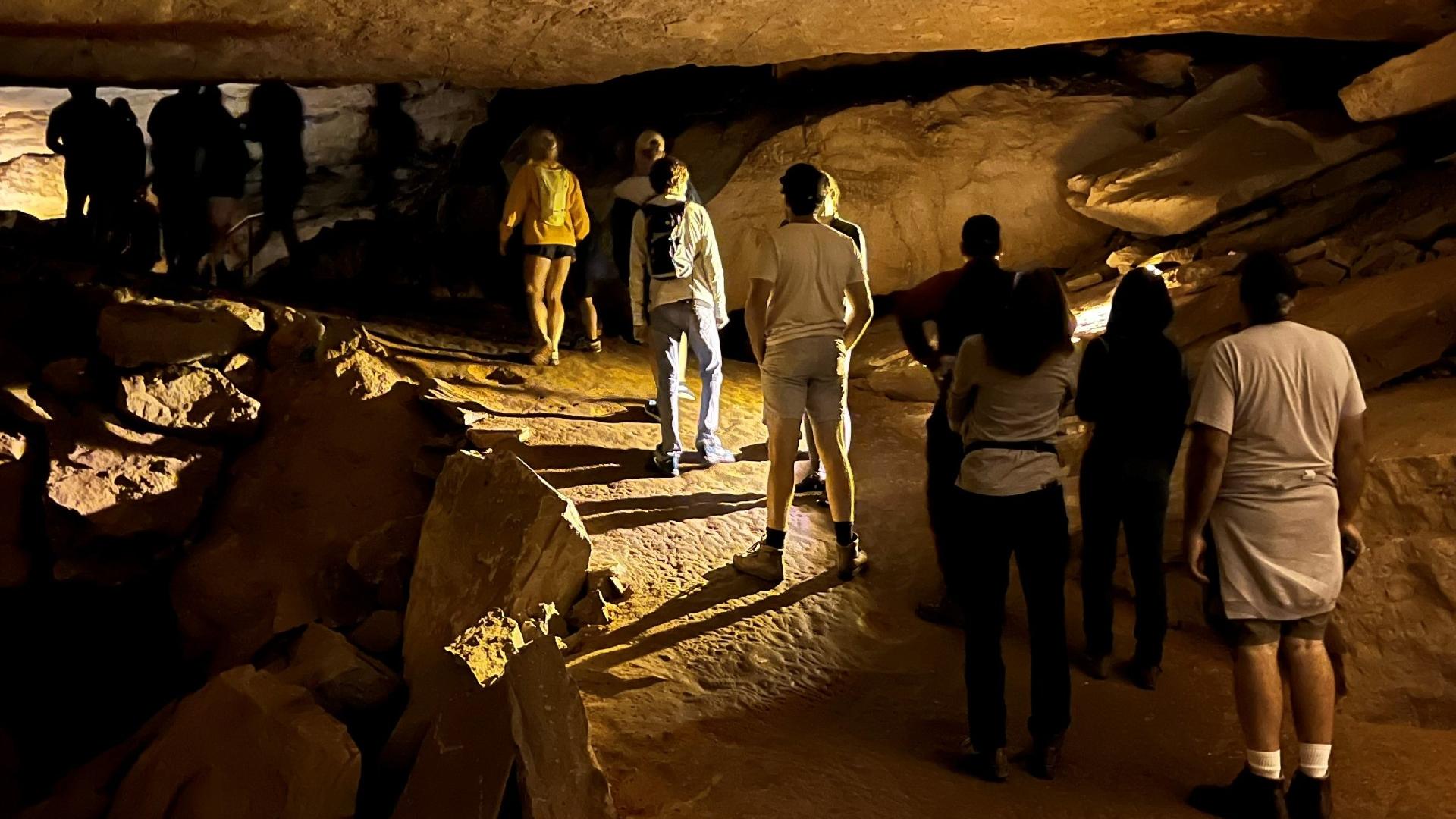 A dozen people stand in a line on a bumpy dirt trail in a dimly lit rock cave.