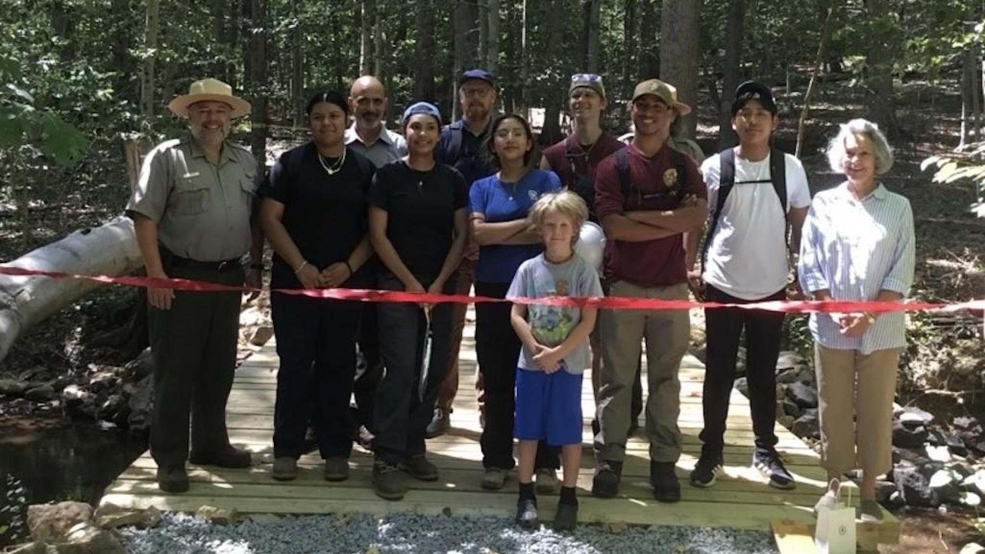 Twelve people standing on wooden bridge in the forest getting ready to cut the red ribbon to open the bridge to visitors.
