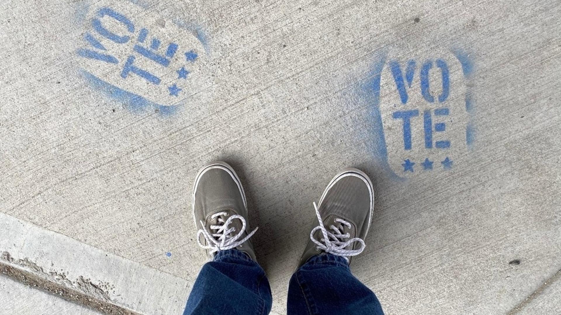 Person in jeans and grey sneakers standing on sidewalk that has been painted with the word "Vote"