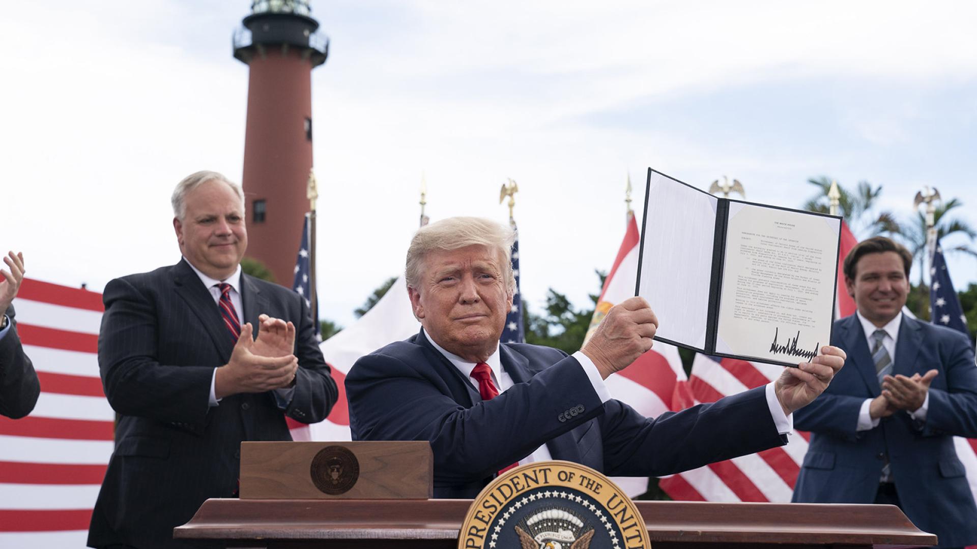 President Trump holds up a signed document while sitting at a table outside with Secretary Bernhardt nearby and a lighthouse in the background.