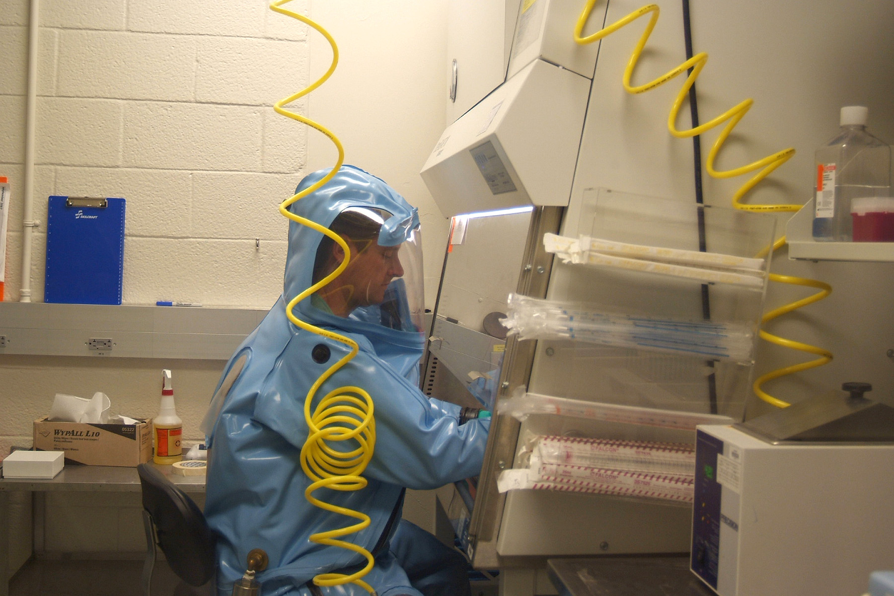 A man at work in a blue full body hazmat suit is surrounded by laboratory equipment