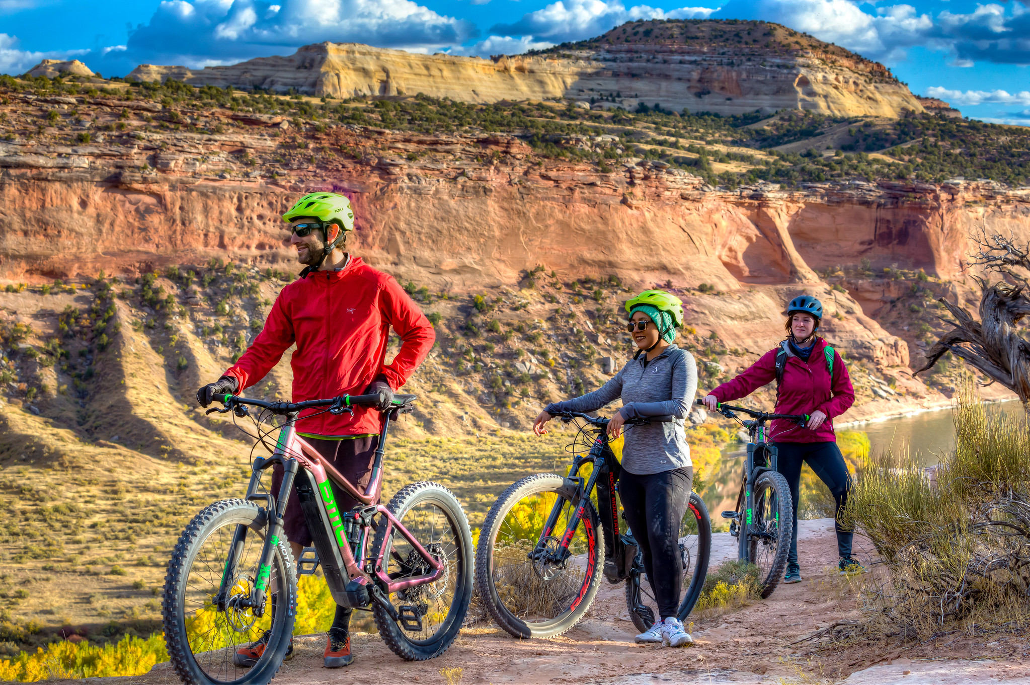 Three people wearing helmets and riding gear stand next to bikes on a dirt trail with a large mesa in the background.
