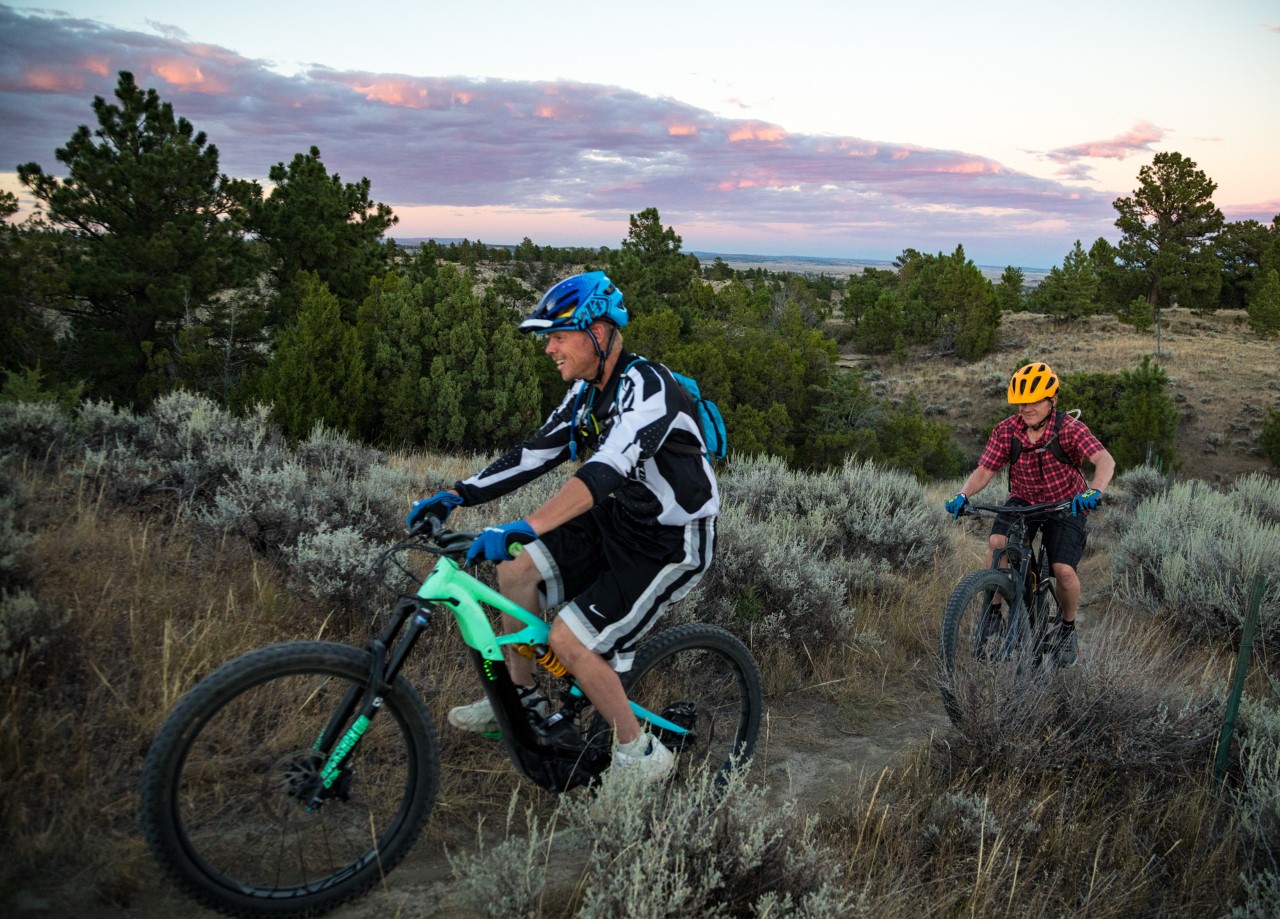 Two people in helmets ride bikes on a narrow dirt trail across a grassy hill near small trees.