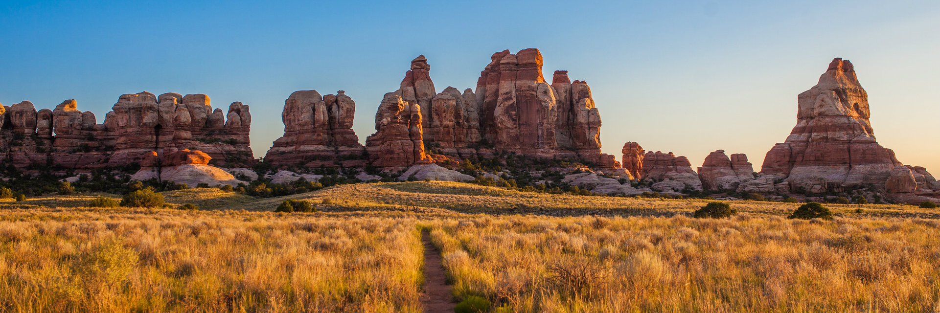 Layered rock formations stick up into a bright blue sky. A path through a glowing yellow field leads straight to them.