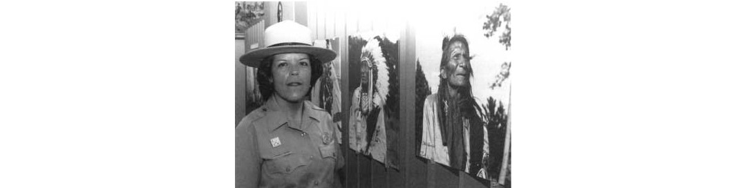 Booher is shown in front of an exhibit displaying photographs of Native Americans. She is wearing the National Park Service 75th Anniversary pin.