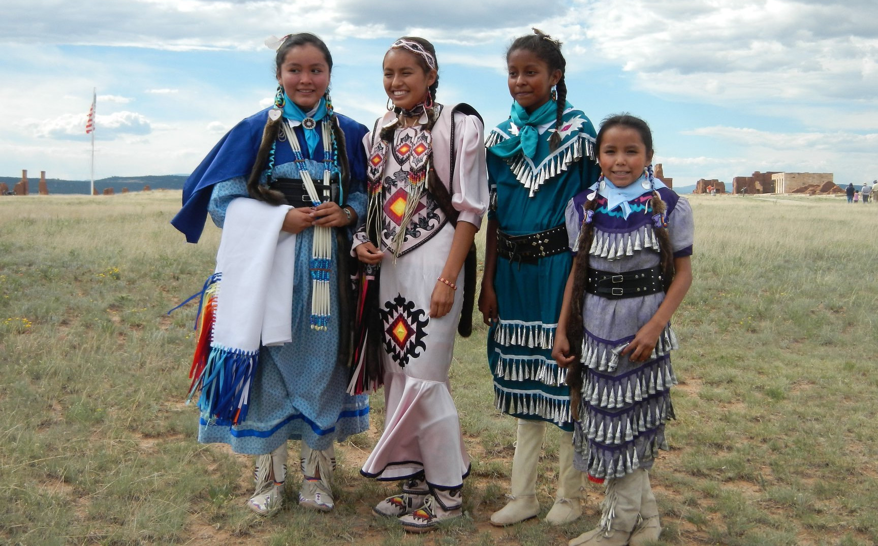 Four young girls in traditional Native American clothes pose together in grassy field.