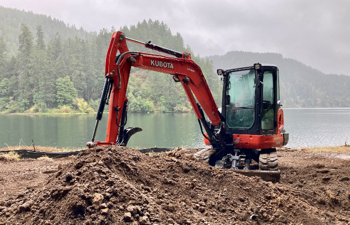 Red construction machine digs brown dirt in front of a foggy lakeside scene with evergreen trees in background