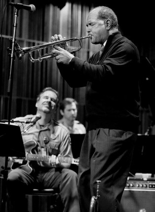 An African American man plays a trumpet while a park ranger playing a guitar sits behind him.