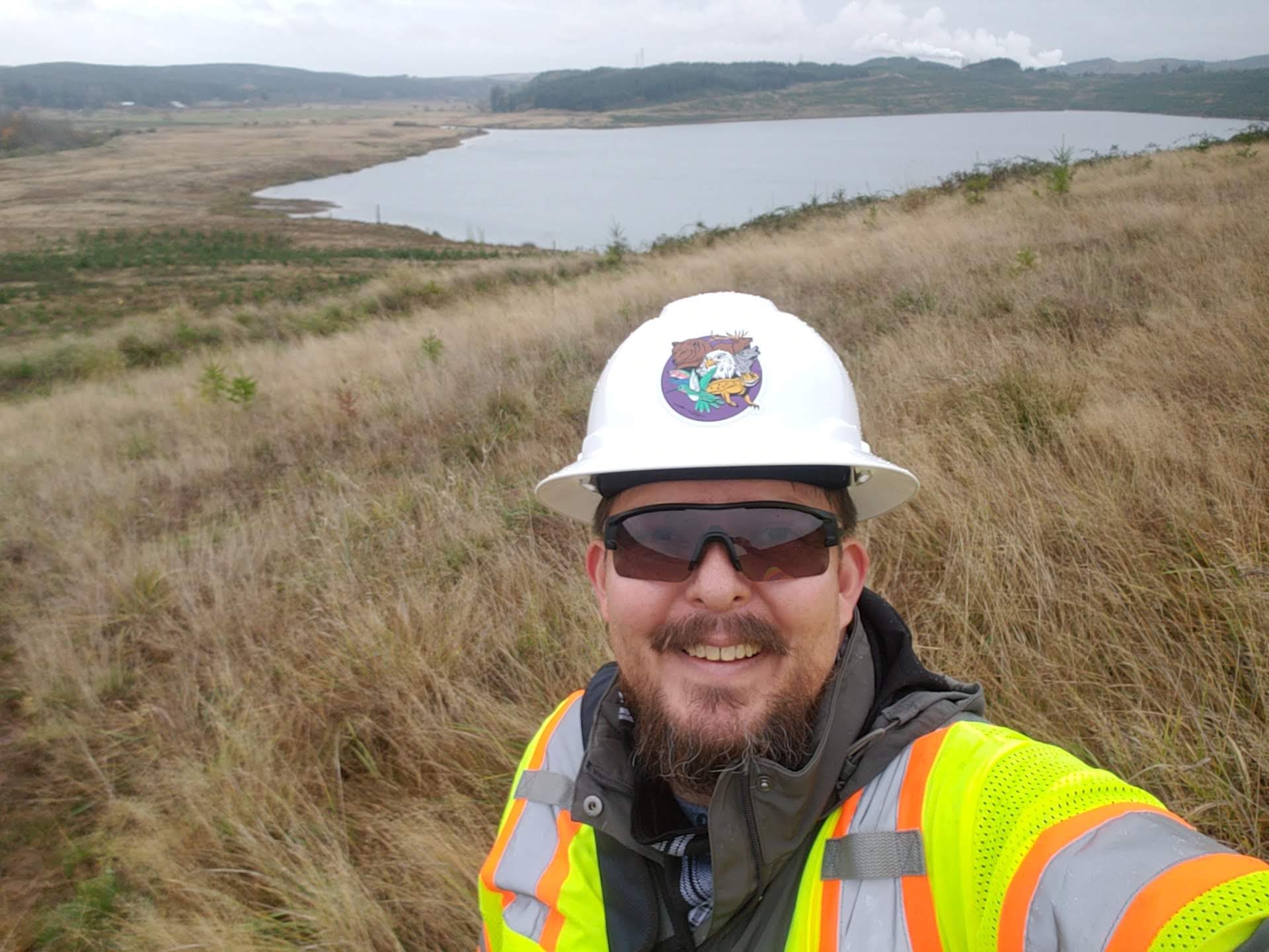 Mychal wearing a hard hat and safety vest, in front of a lake. 