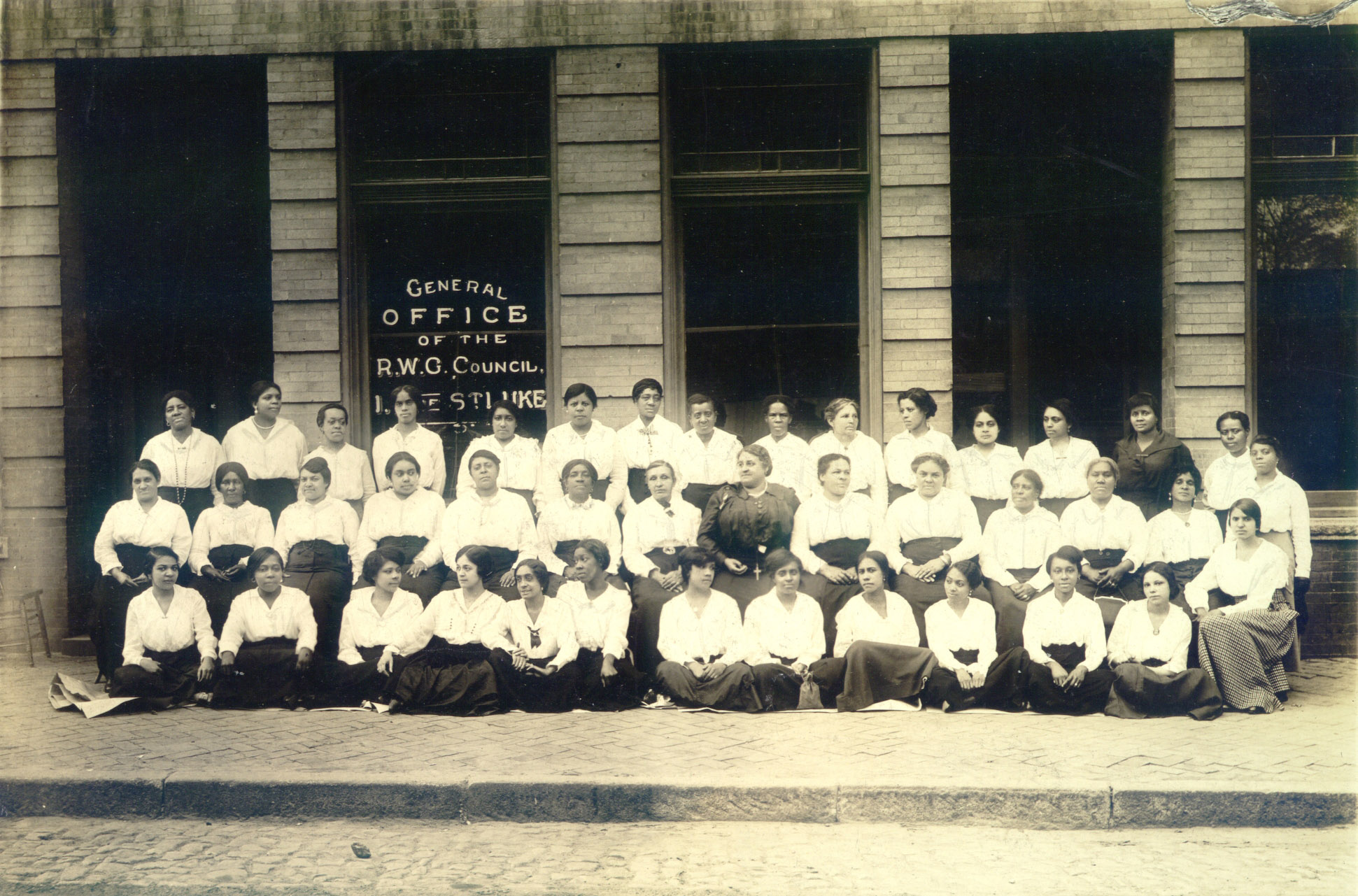 A historic black and white photo of a group of about 50 African American women posing in front of a brick building with large windows.