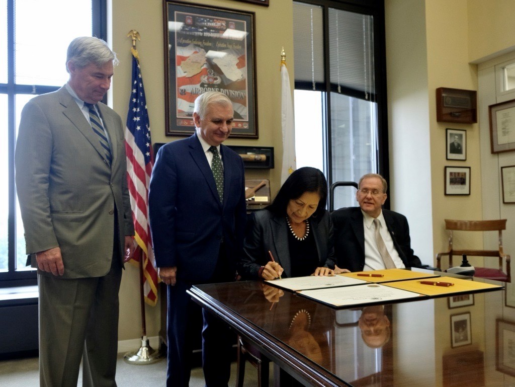 Secretary Haaland signing a document at a desk with members of the Rhode Island congressional delegation.