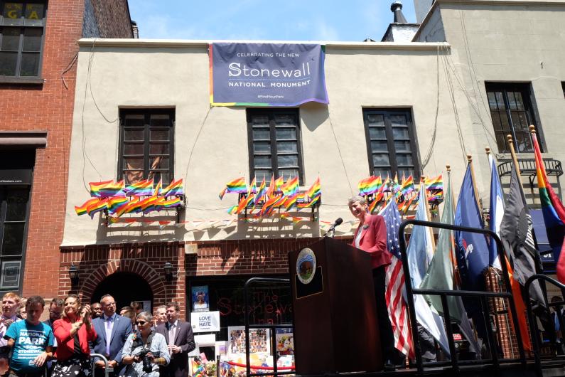 Secretary Jewell addresses a crowd while standing behind a wooden podium and in front of a chorus of rainbow flags.