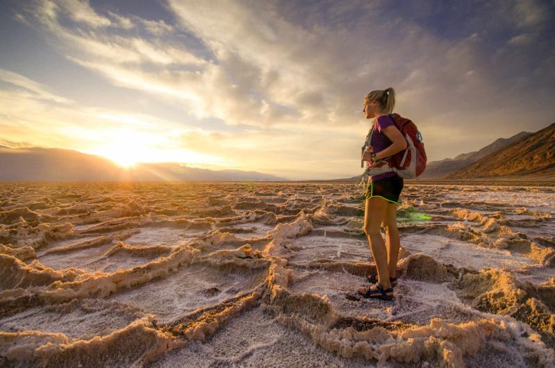 A young woman stands on the desert floor among strange salt formations watching the sun set.