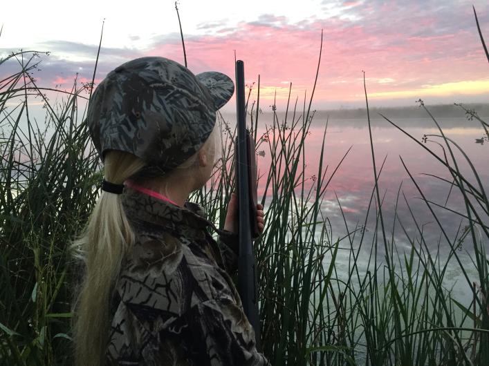 Hunting at Minnesota Valley National Wildlife Refuge in Minnesota. A young girl with blonde hair wears camouflage hunting clothes and holds a shotgun while standing in tall grass and looking at a pink sunrise.