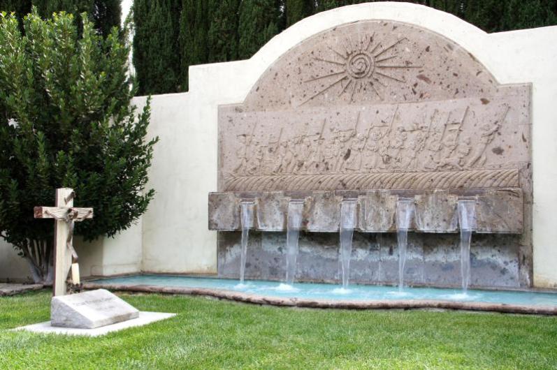 A small cross tombstone stands in a grassy courtyard next to a flowing fountain.